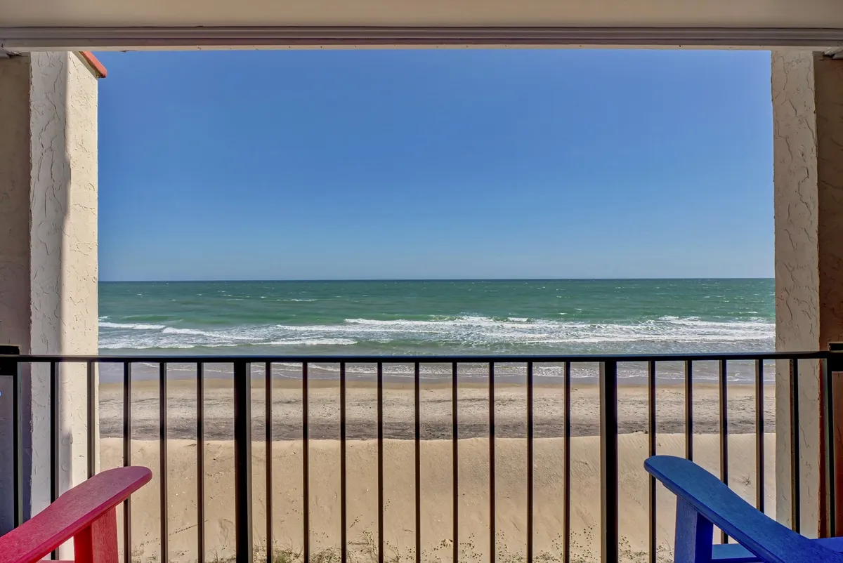 This image features a private condo balcony overlooking the rolling ocean waves and a wide stretch of sandy beach. Colorful chairs are arranged for comfort, suggesting a space for relaxing, reading, or socializing while enjoying serene coastal views. The tranquil mood is enhanced by the open sky and the inviting expanse of the shoreline below.
