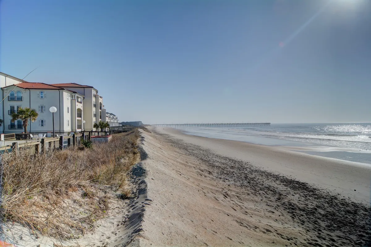 A scenic view of beachfront properties, with a wooden pier extending into rolling ocean waves. The scene invites guests to walk along the sand, fish from the pier, or simply soak in the coastal ambiance.