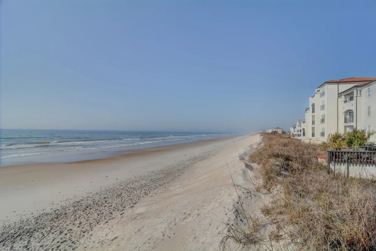 A serene beachfront view with gentle ocean waves, sandy dunes, and coastal vegetation, framed by nearby buildings. The scene captures the easy transition from the property to the beach, inviting families to enjoy sandcastle building, beach games, and seaside walks.