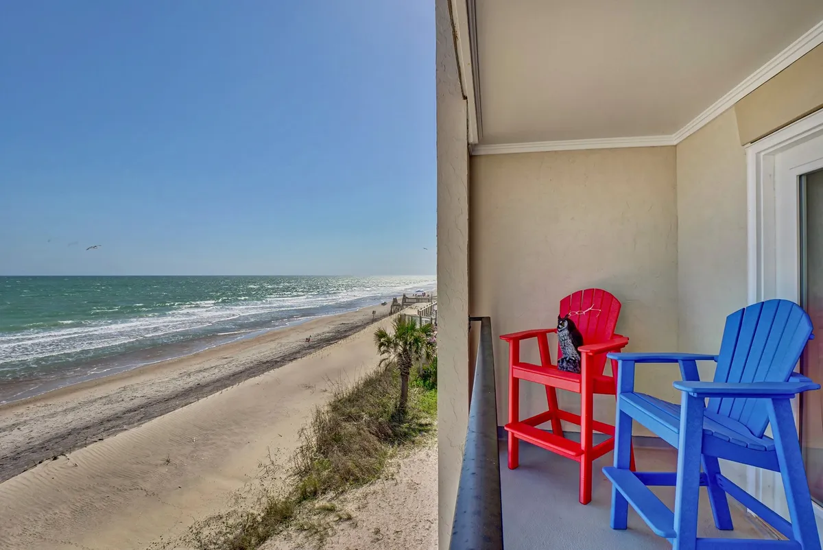 Brightly colored chairs are arranged on a deck or terrace overlooking a sweeping ocean view. The sandy beach and sparkling water are visible just beyond, creating a perfect spot to relax, read, or simply enjoy the scenery.