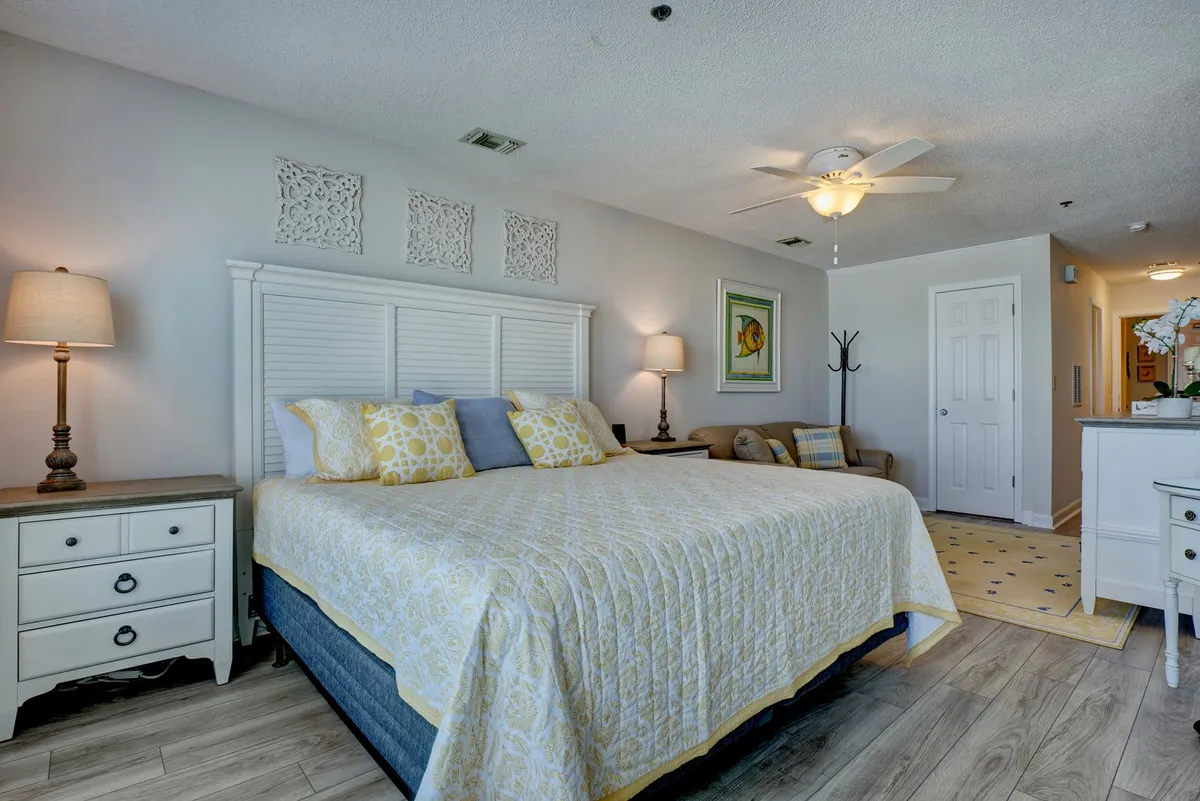 This image features a king-sized bed adorned with plush yellow and gray pillows, complemented by decorative wall pieces, a bedside lamp, and a vibrant fish artwork. Natural light brightens the cozy, relaxing atmosphere, highlighting the room’s inviting textures and bold-yet-soothing color palette.