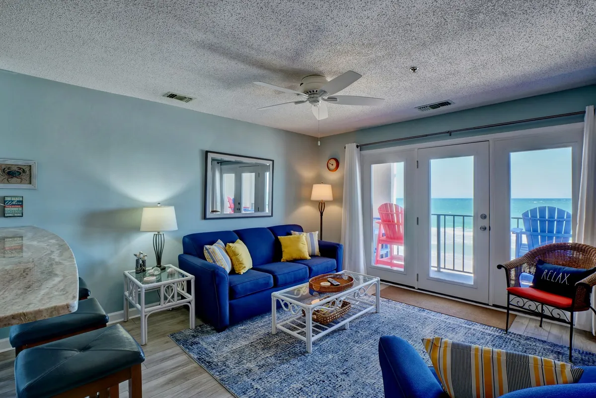 A cozy living area with a blue sofa, complemented by red chairs visible on the terrace beyond. The coffee table is thoughtfully decorated, and the sliding doors open up to reveal the terrace and a stunning ocean view, reinforcing the connection between indoor comfort and outdoor beauty.