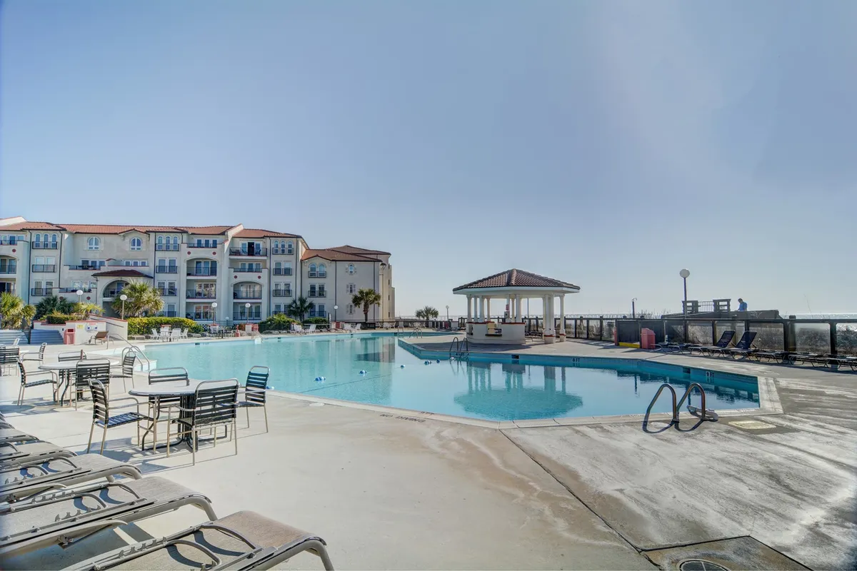 A lively exterior scene featuring a large swimming pool surrounded by numerous lounge chairs and a charming gazebo, with the villa's apartment building as a backdrop. The area is designed for both relaxation and socialization, inviting guests to swim, sunbathe, or gather with friends by the water.