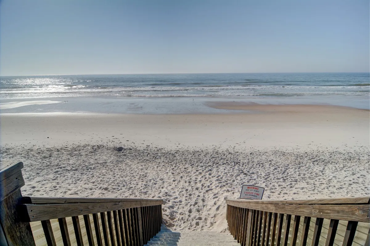 Wooden stairs descend to a wide, sandy beach bordered by calm ocean waves and a clear blue sky. The inviting beach is just steps from the resort, emphasizing effortless access to sun, sand, and surf.