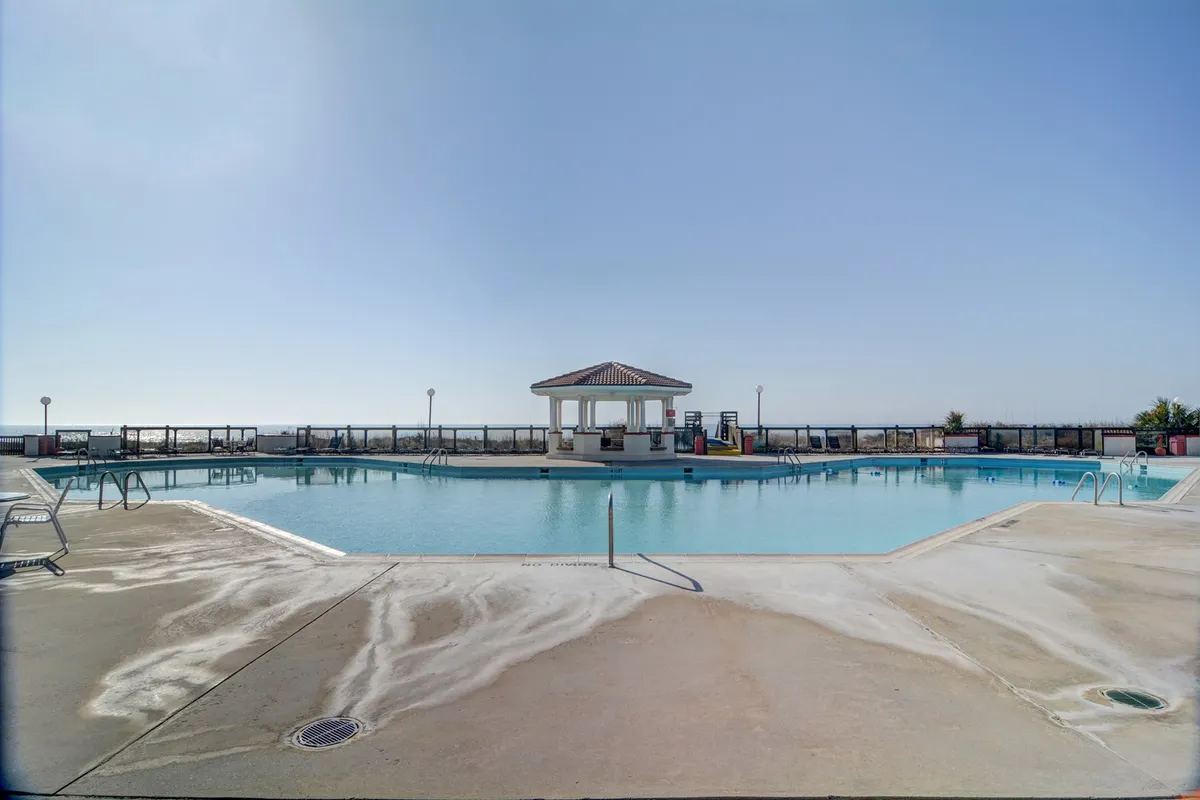 A vibrant outdoor pool area framed by lush landscaping and a pool house, with an array of lounge chairs set up for sunbathing or relaxing. The scene suggests an inviting atmosphere where families can swim, gather, and unwind under the open sky.