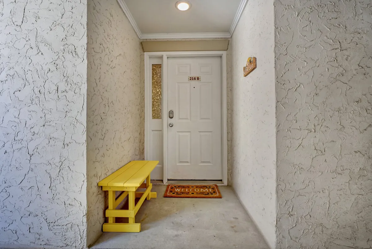 A welcoming entryway with a bright yellow bench, a 'welcome' mat, and the front door, all presented in a cheerful, family-friendly style. The area feels inviting and sets a positive first impression for guests arriving at the villa.
