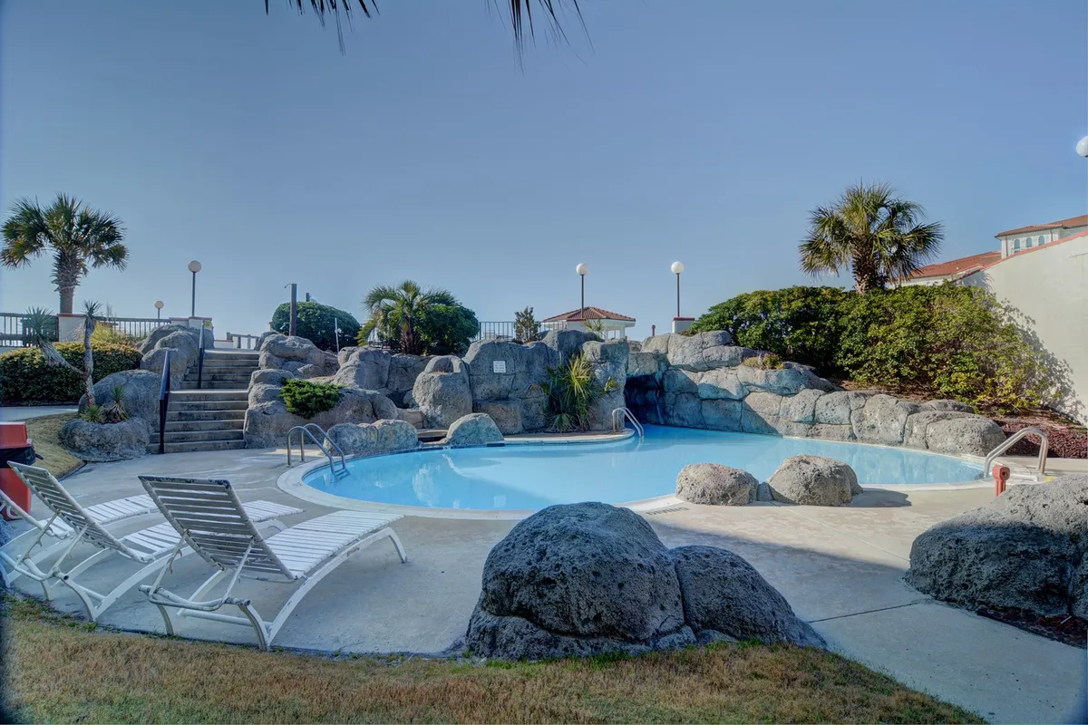 An inviting outdoor pool area with crystal-clear water, rock formations for added character, and plenty of lounge chairs. Landscaped greenery surrounds the scene, creating a tranquil yet social atmosphere ideal for unwinding or meeting new friends.
