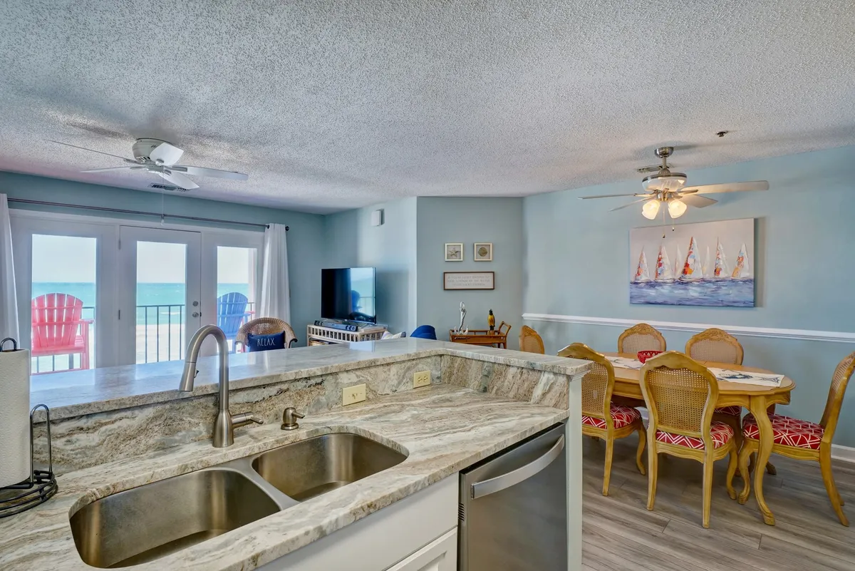 This interior image captures a spacious kitchen island with a sink, a welcoming dining table, and a panoramic ocean view through the balcony doors. The colorful chairs on the balcony hint at the coastal setting and invite relaxation just beyond the dining area. The scene radiates warmth, family togetherness, and the enjoyment of meals enhanced by stunning scenery.