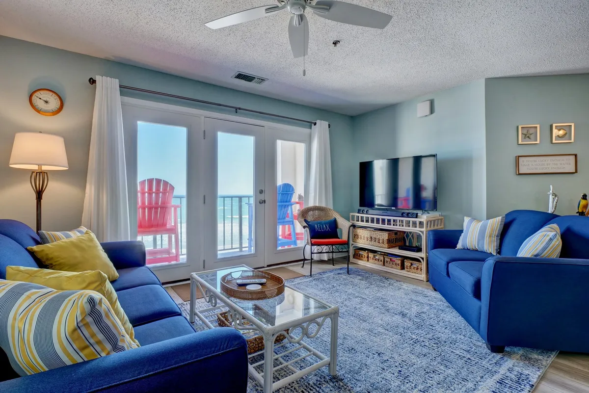 A spacious living room featuring plush blue sofas, a glass coffee table, and a large TV. Sunlight streams through sliding glass doors that open directly to a private terrace, offering panoramic ocean views. The décor is vibrant yet calming, with beach-themed accents and an open layout that encourages relaxation and gatherings.