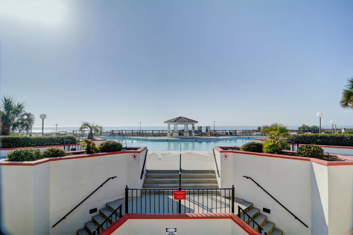 A vibrant exterior shot of the resort’s oceanfront swimming pool, framed by lush palm trees and a pavilion. Lounge chairs line the poolside, inviting relaxation and sunbathing in a tropical setting just steps from the beach.